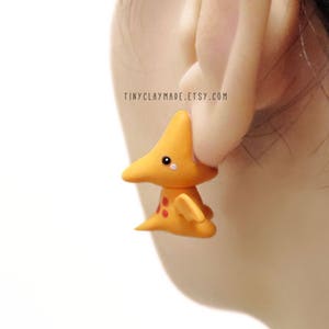 May include: A pair of yellow clay earrings shaped like a cartoon dinosaur with red spots. The earrings are worn by a person with dark hair.