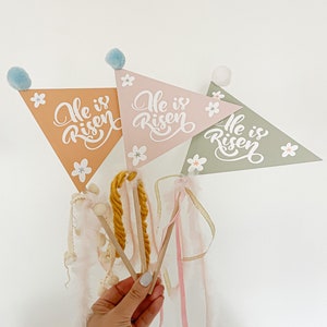 Pode incluir: Três bandeiras triangulares de Páscoa em tons de pêssego, rosa e verde sálvia, cada uma com o texto "He is Risen" e detalhes florais brancos. As bandeiras têm varetas de madeira, pompons e fitas. Uma mão segura as bandeiras contra um fundo branco.