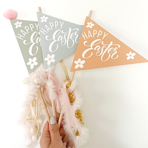 May include: Triangular Easter banners in sage green, light blue, and peach, each with "Happy Easter" in white script and white flower accents. The banners are attached to wooden dowels and adorned with pink feathers and ribbons.