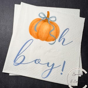 May include: White paper napkins with an orange pumpkin illustration and blue ribbon. The napkins have the words "sh boy!" written in blue script. The napkins are stacked on top of each other.