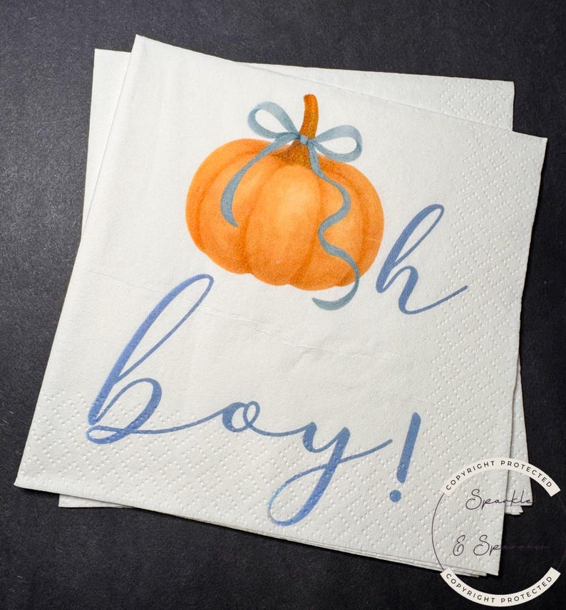 May include: White paper napkins with an orange pumpkin illustration and blue ribbon. The napkins have the words "sh boy!" written in blue script. The napkins are stacked on top of each other.
