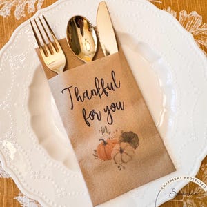 May include: A gold fork, spoon, and knife set inside a brown paper sleeve on a white plate. The sleeve has the text "Thankful for you" and a pumpkin illustration. The plate is on a yellow floral tablecloth.