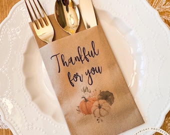 May include: A brown paper cutlery sleeve containing a gold fork, spoon, and knife. The sleeve features the black text Thankful for you and a printed illustration of three pumpkins in orange, beige, and dark green colors on a white plate.