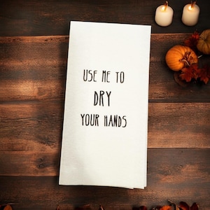 May include: White kitchen towel with black text that reads "Use me to dry your hands". The towel is on a brown wooden surface with fall decorations.