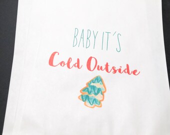 May include: White paper gift bag with the text "Baby It's Cold Outside" in teal and red lettering. A stylized green and orange Christmas tree is printed below the text.