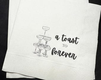 May include: White paper napkins with a black illustration of champagne glasses stacked in a pyramid. The text "a toast to forever" is printed on the napkins. The napkins are stacked on a black surface.