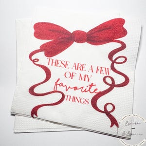 May include: White paper napkins with a red bow and ribbon design. The text "THESE ARE A FEW OF MY favorite THINGS" is printed in red. The napkins are stacked on top of each other.