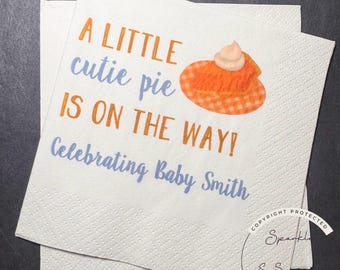 May include: Two white paper napkins with the text "A LITTLE cutie pie IS ON THE WAY! Celebrating Baby Smith" in orange and blue. A cartoon pumpkin pie graphic is also present.