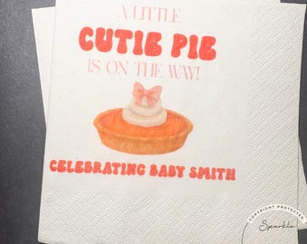 May include: White paper napkins with a baby shower theme. The napkins feature the text "A LITTLE CUTIE PIE IS ON THE WAY!" and "CELEBRATING BABY SMITH" in orange. A pie graphic with whipped cream and a bow is also present.