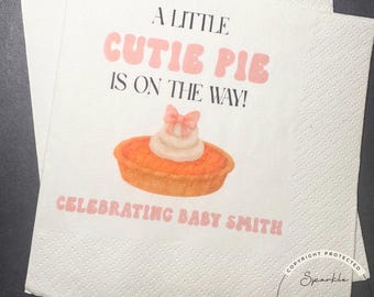 May include: White paper napkins with the text "A LITTLE CUTIE PIE IS ON THE WAY!" and "CELEBRATING BABY SMITH". The napkins feature a watercolor illustration of a pie with whipped cream and a bow.