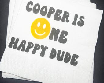 May include: A stack of white paper napkins with the text "COOPER IS ONE HAPPY DUDE" in black, with a yellow smiley face. The napkins are stacked on a dark surface.