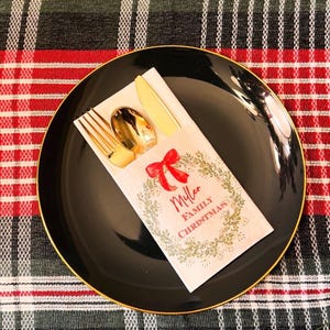 May include: A black plate with a gold rim holds a napkin with a red bow and the words "Miller Family Christmas." Gold-colored fork, spoon, and knife are inside the napkin. The plate rests on a red and black plaid tablecloth.
