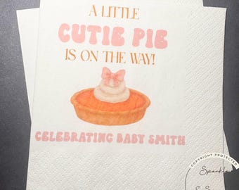 May include: White paper napkins with the text "A LITTLE CUTIE PIE IS ON THE WAY!" and "CELEBRATING BABY SMITH" in pink. The napkins feature a watercolor illustration of a pie with whipped cream and a bow.