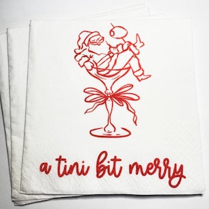 May include: A stack of white paper napkins featuring a red line art illustration of Santa Claus relaxing inside a martini glass with an olive. Below the illustration, the text a tini bit merry is printed in a red cursive script font.
