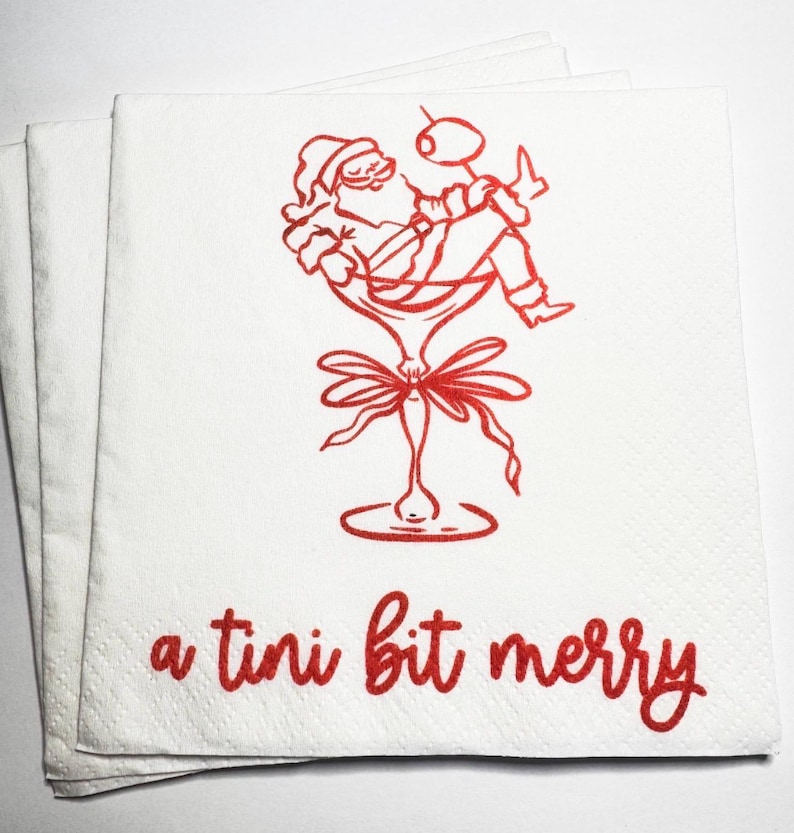 May include: A stack of white paper napkins featuring a red line art illustration of Santa Claus relaxing inside a martini glass with an olive. Below the illustration, the text a tini bit merry is printed in a red cursive script font.