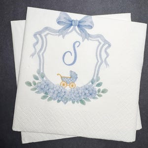 May include: White paper napkins with a blue watercolor design. The design includes a blue bow, a shield with the letter "S", a baby carriage, and blue flowers. The napkins are stacked on top of each other.