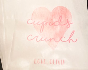 May include: White paper bags with a pink watercolor heart design. The heart has the text "cupid's crunch" in pink script. The bottom of the bag has the text "LOVE, OLIVIA" in pink script.