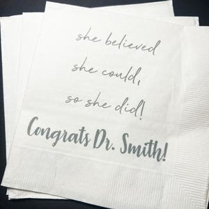 May include: White paper napkins with the text "she believed she could, so she did! Congrats Dr. Smith!" in a silver script font. The napkins are stacked and have a textured surface.