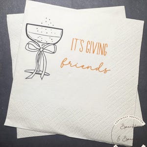 May include: Two white paper napkins with a textured pattern. One napkin is on top of the other. The top napkin has a black and white drawing of a champagne glass with a bow and the words "IT'S GIVING friends" in orange.