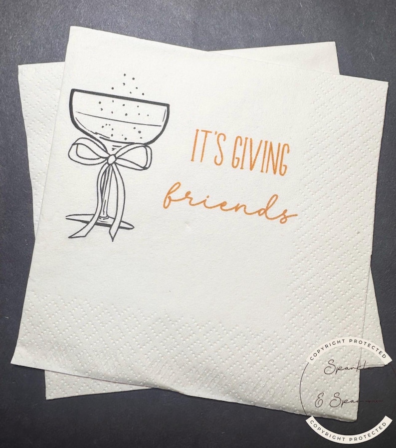 May include: Two white paper napkins with a textured pattern. One napkin is on top of the other. The top napkin has a black and white drawing of a champagne glass with a bow and the words "IT'S GIVING friends" in orange.