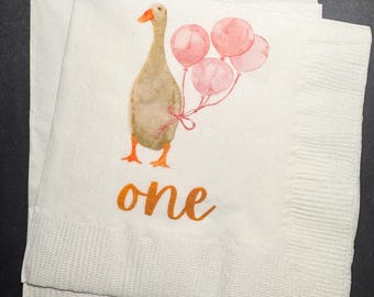 May include: White paper napkins with a watercolor illustration of a goose holding pink balloons. The word "one" is written in orange script below the goose. The napkins are folded and stacked.