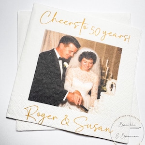 May include: A white square paper napkin featuring a vintage black and white photograph of a couple cutting a wedding cake. The text "Cheers to 50 years!" is printed in gold script at the top, and "Roger & Susan" is printed in gold script at the bottom.