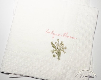 Op de afbeelding: Witte papieren servetten met een bloemenboeket illustratie en de tekst "baby in bloom" in roze script. De servetten hebben een getextureerd oppervlak en zijn opgestapeld. Een copyrightteken staat in de rechter benedenhoek.