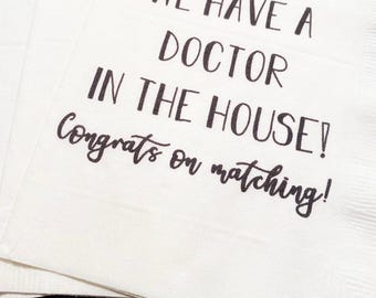May include: White paper napkins with black text that reads "WE HAVE A DOCTOR IN THE HOUSE! Congrats on matching!" The napkins are stacked and slightly fanned out.