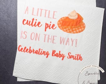 May include: White paper napkins with the text "A LITTLE cutie pie IS ON THE WAY! Celebrating Baby Smith" in pink and red. A pie graphic is also included. The napkins are for a baby shower.