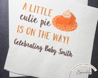 May include: White paper napkins with the text "A LITTLE cutie pie IS ON THE WAY! Celebrating Baby Smith" in brown and orange lettering. A pumpkin pie graphic is also featured on the napkins. The napkins are for a baby shower.