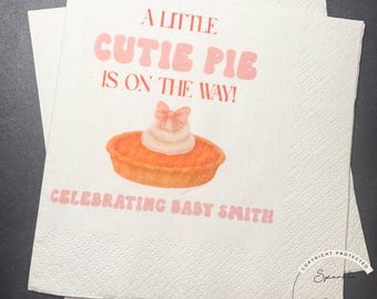 May include: White paper napkins with the text "A Little Cutie Pie Is On The Way!" in pink. A pie graphic with a bow is in the center. The text "Celebrating Baby Smith" is below the pie graphic.