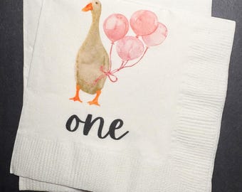 May include: White paper napkins with a watercolor illustration of a goose holding pink balloons. The word "one" is written in black script below the goose. The napkins are folded and stacked.