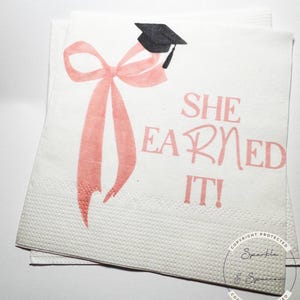 May include: White paper napkins with a pink bow and graduation cap design. The text "SHE EARNED IT!" is printed in pink. The napkins are stacked and have a textured surface, suitable for a graduation celebration.