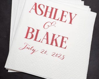 May include: A stack of white paper napkins with red glitter text. The text reads "ASHLEY & BLAKE" above "July 20, 2025". The napkins have a subtle, textured pattern and are arranged on a dark surface.