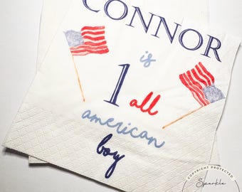 Può includere: Tovaglioli di carta bianchi con motivo a rombi. I tovaglioli sono decorati con la scritta "CONNOR is 1 all american boy" in blu e rosso, insieme a piccole bandiere americane.