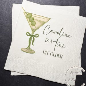 May include: White cocktail napkins with a martini glass illustration and the text "Caroline is a tini bit older". The martini glass has green olives and a green bow.