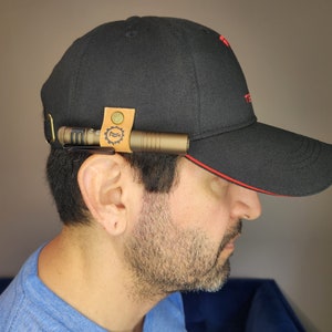 May include: A black baseball cap with a red brim and a leather strap with a metal clip to hold a flashlight. The flashlight is a brown metal color.