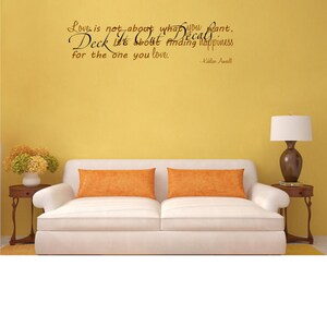Puede incluir: Un sofá blanco con dos almohadas naranjas en una sala de estar con una pared amarilla. La pared tiene una calcomanía de vinilo negro con la cita "El amor no se trata de lo que quieres. Deck se trata de encontrar la felicidad para la persona que amas. -Kahlan Amnell"