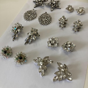 Op de afbeelding: Een verzameling zilverkleurige strass oorbellen in diverse ontwerpen, waaronder sterren, bloemen en druppels. De oorbellen zijn versierd met heldere, sprankelende strass-steentjes, sommige met parels. Ze liggen op een witte ondergrond.