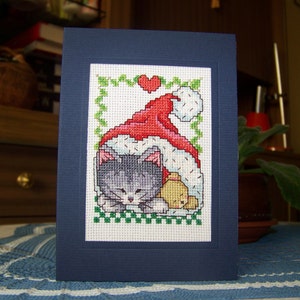 Handgemaakte kerstkaart kat in hoed met kruissteek. Feestdagen