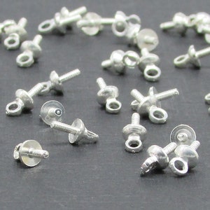 May include: A collection of small, silver-colored jewelry findings, including screw posts, eye pins, and flat head pins. These components are scattered across a dark gray surface, showcasing their intricate designs and potential for jewelry making.