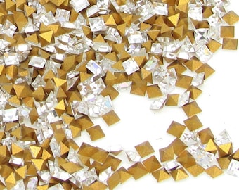 May include: A pile of gold and clear glass pyramid-shaped rhinestones. The rhinestones are scattered on a white background.