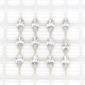 May include: Twelve clear, marquise-cut gemstones arranged in a grid pattern. Each gem has a pointed tip at both ends, with facets catching the light. The gems are set against a white, square-patterned background.
