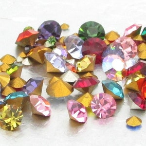 May include: Assortment of colorful rhinestones in various shapes and sizes. The rhinestones are scattered on a white surface.