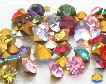 May include: Assortment of colorful rhinestones in various shapes and sizes. The rhinestones are scattered on a white surface.