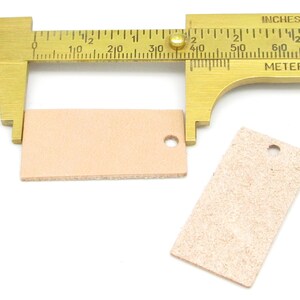 May include: Two light brown leather tags with a hole punched in the top. A brass measuring tool is measuring the tag on the left, showing it is 1 3/8 inches wide.