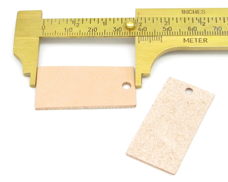 May include: Two light brown leather tags with a hole punched in the top. A brass measuring tool is measuring the tag on the left, showing it is 1 3/8 inches wide.