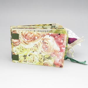 May include: A handmade journal featuring a rose floral design on the cover. The journal is bound with green fabric and has a decorative tag with a purple accent. The pages are visible and appear to be made from recycled paper.