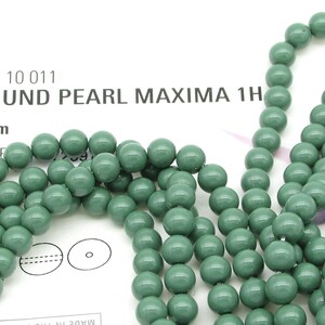 Pu&ograve; includere: Un filo di perle rotonde verdi da 6 mm. Le perle hanno una finitura opaca e una leggera variazione di colore. Le perle sono infilate su un filo bianco. L'immagine include anche il testo "131 10 011 ROUND PEARL MAXIMA 1H 6 mm SAGE"