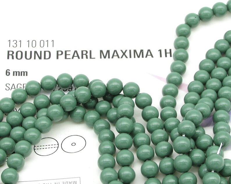 Pu&ograve; includere: Un filo di perle rotonde verdi da 6 mm. Le perle hanno una finitura opaca e una leggera variazione di colore. Le perle sono infilate su un filo bianco. L'immagine include anche il testo "131 10 011 ROUND PEARL MAXIMA 1H 6 mm SAGE"